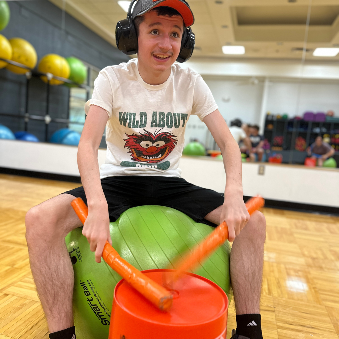 A participant is in a fitness studio, seated on a green medicine ball. They are drumming with foam sticks on a reverse bucket while wearing headphones. The participant is smiling.