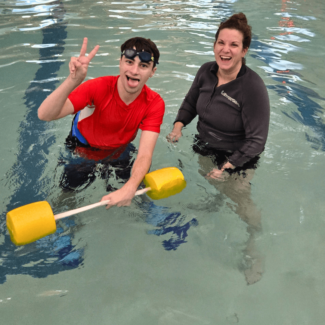 An image of a Rec2Connect Aquatic Specialist with a participant during an aquatic session. The participant is holding a floating barbell and holding up a peace sign.