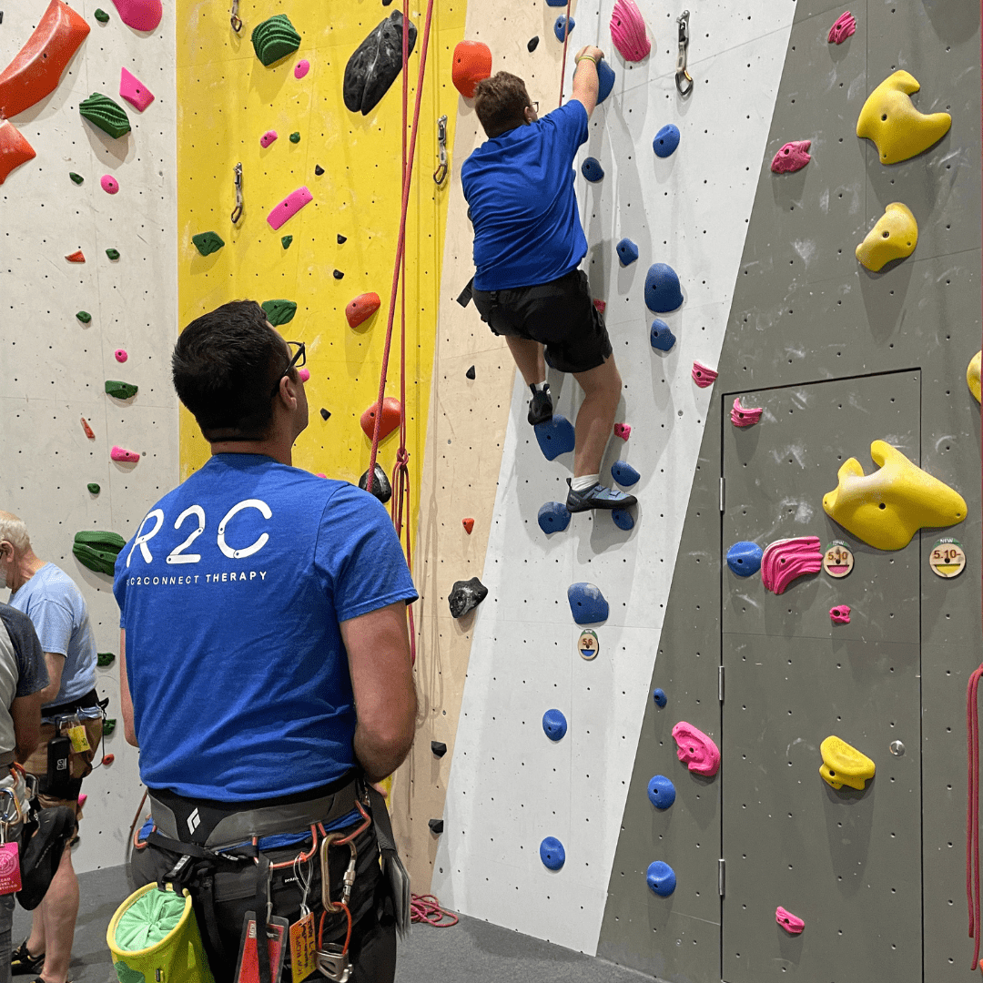A participant is belayed by a volunteer in a colorful indoor rock gym.