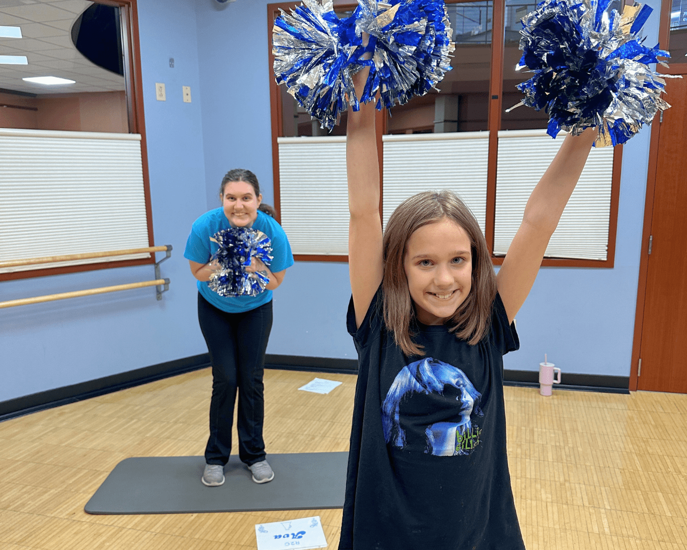 Two participants in a fitness studio, one younger and the other older, are cheering with blue and silver-colored pom poms.
