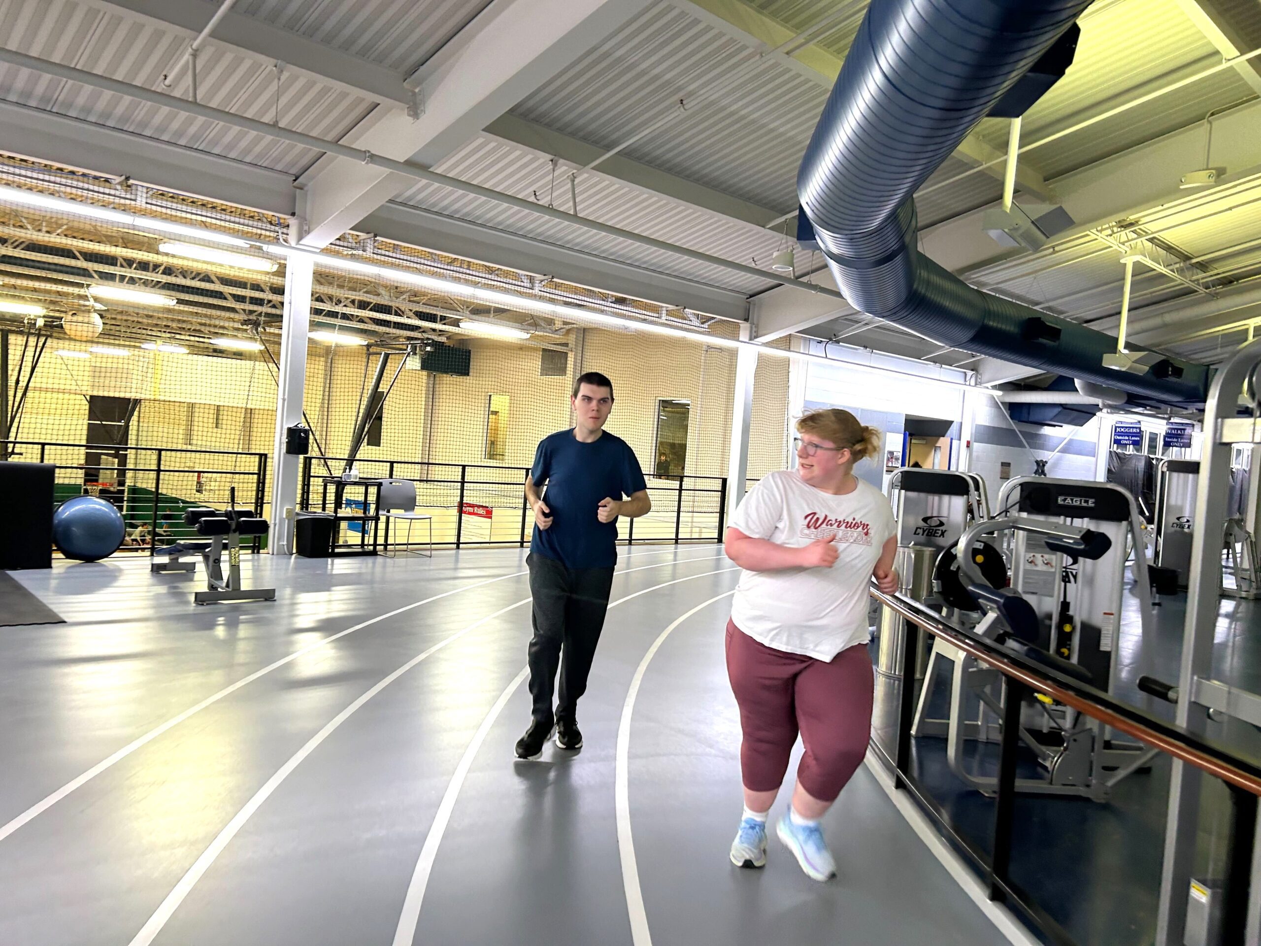 Two participants are running on an indoor track, with gym equipment visible in the background.